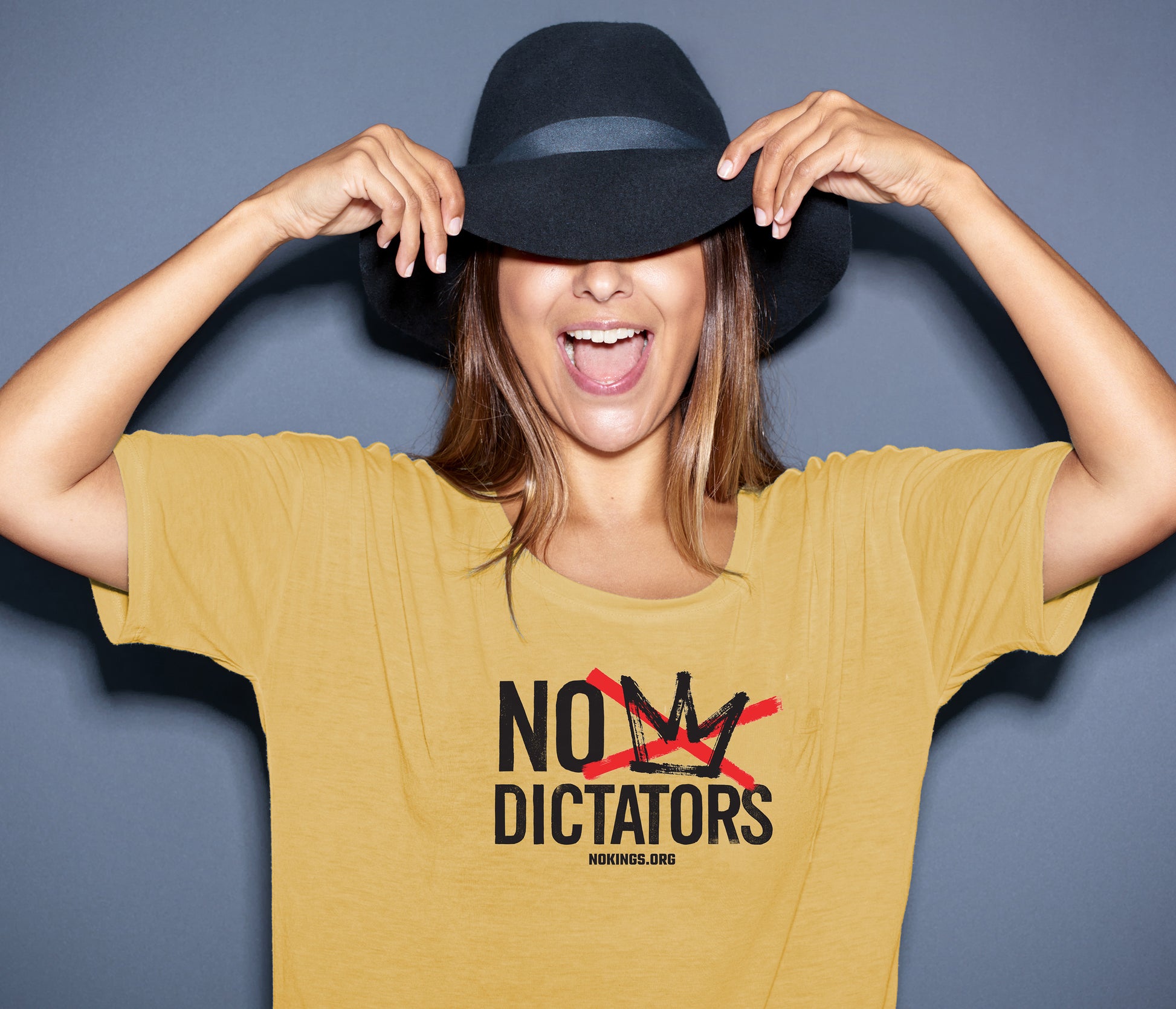 Person wearing a mustard yellow t-shirt with 'No Dictators' text, holding a black hat.